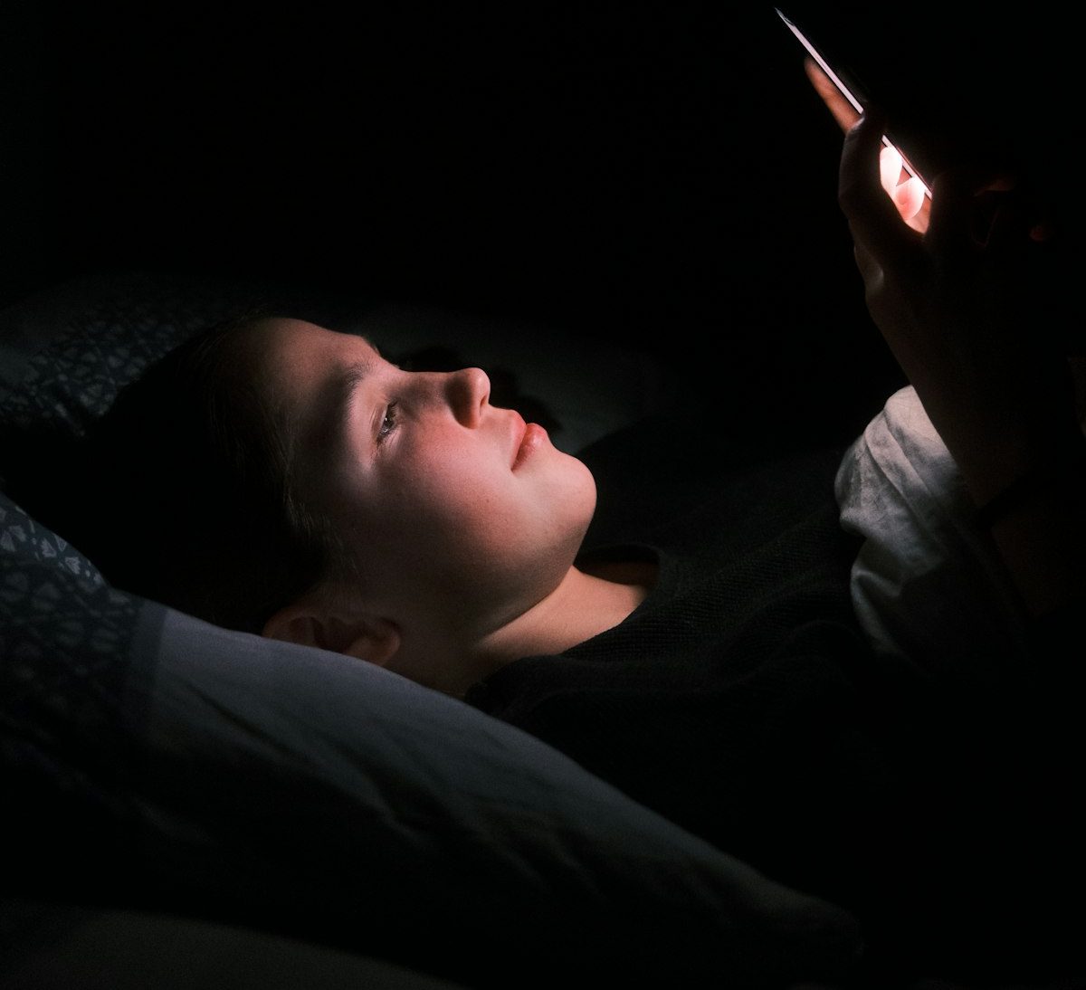 Una chica acostada en la cama de noche, con la cara iluminada por la pantalla del teléfono